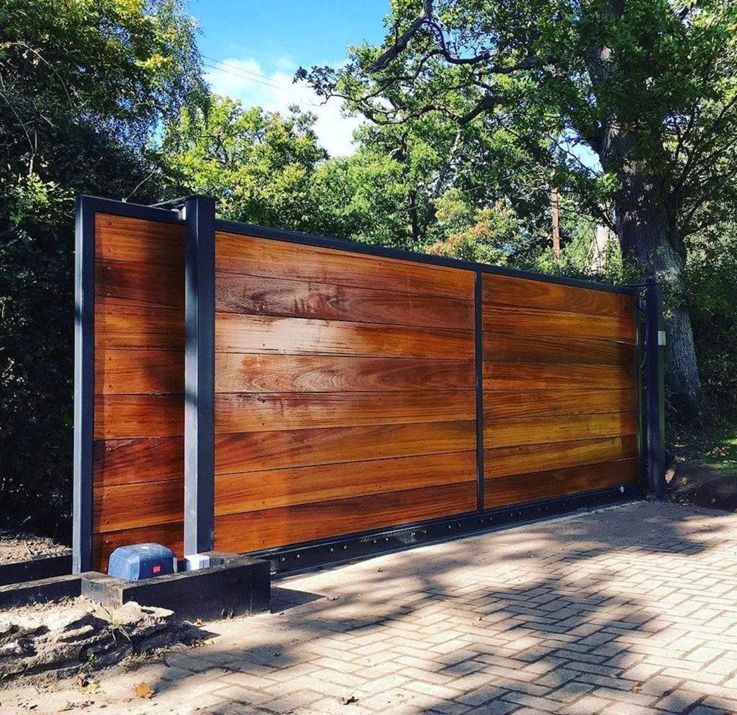 Wide slide gate of horizontal hardwood boards in a dark steel frame across a herringbone brick drive, with the gate operator on a low plinth beside the post and trees behind