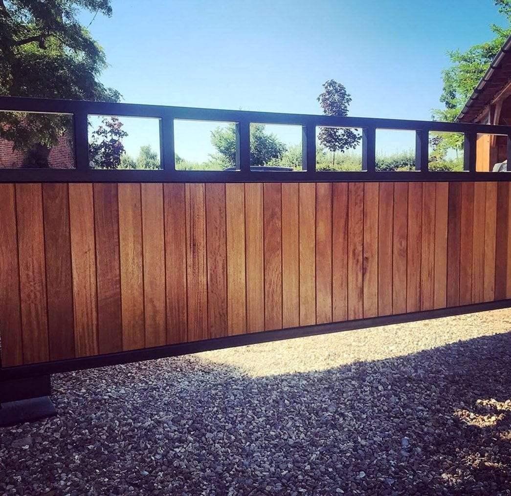 Slide gate of vertical hardwood boards topped by an open steel frame of square panels, standing on a gravel drive in bright sun