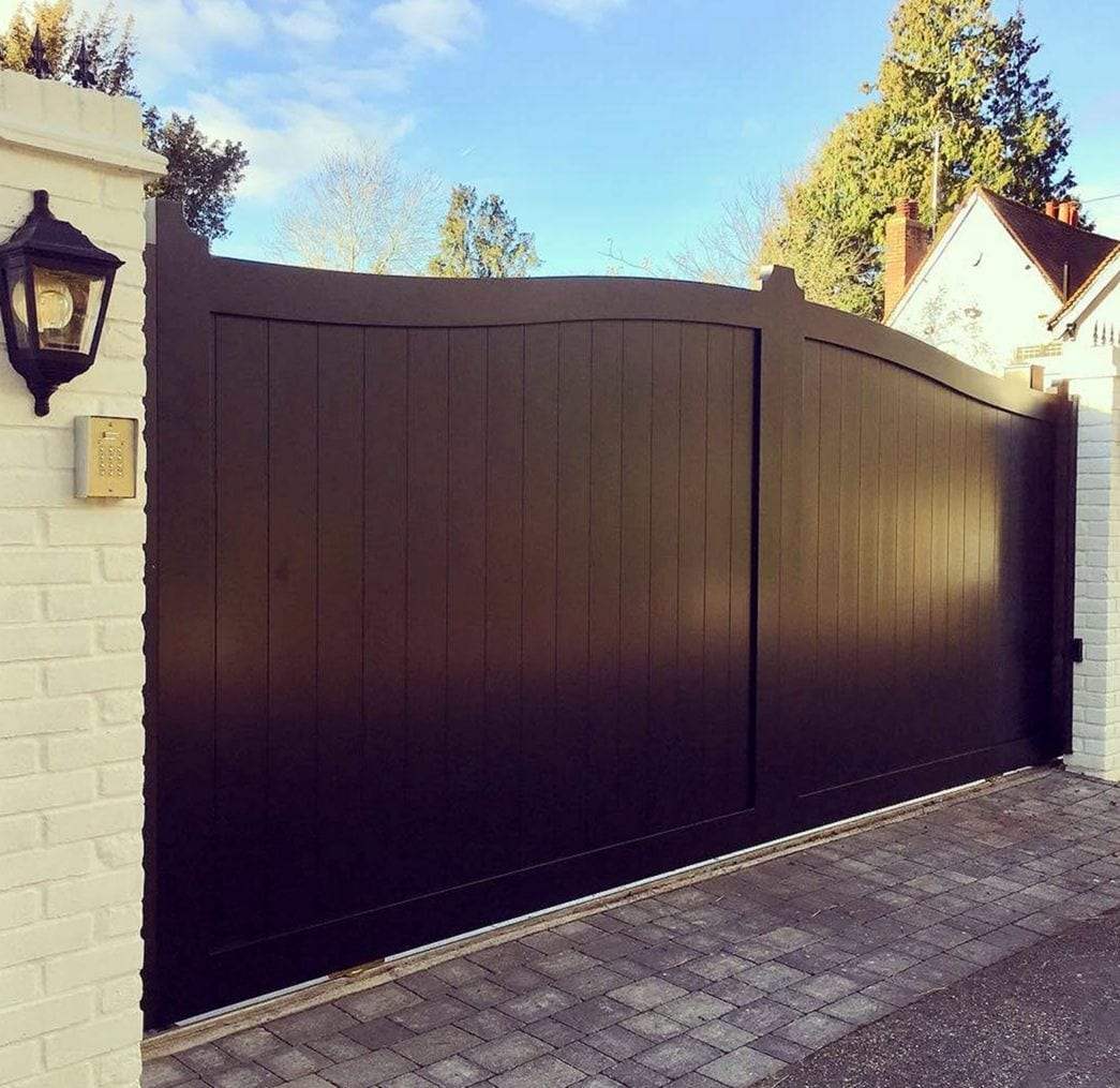Dark solid gate with vertical boarding and a gently curved top, running on a metal track set into block paving between white painted brick piers, with a keypad and a black lantern on the left pier