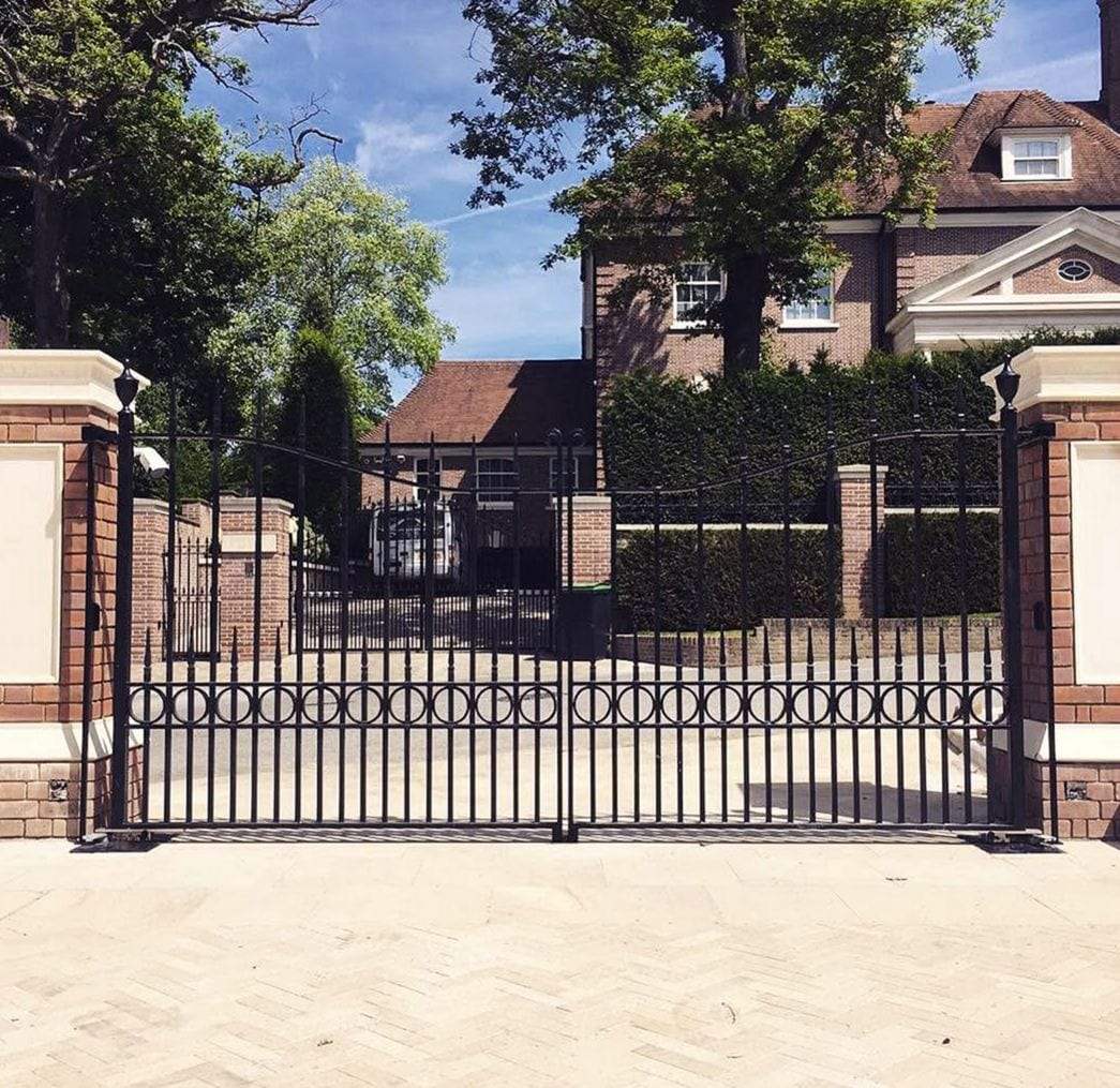Pair of black metal swing gates with spear-top pickets, a decorative band of rings and an arched top rail, hung between brick and stone piers across a pale paved driveway