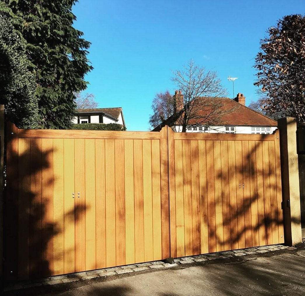 Pair of closed hardwood swing gates with framed vertical boards and a flat top, set between square piers across a driveway under a blue winter sky