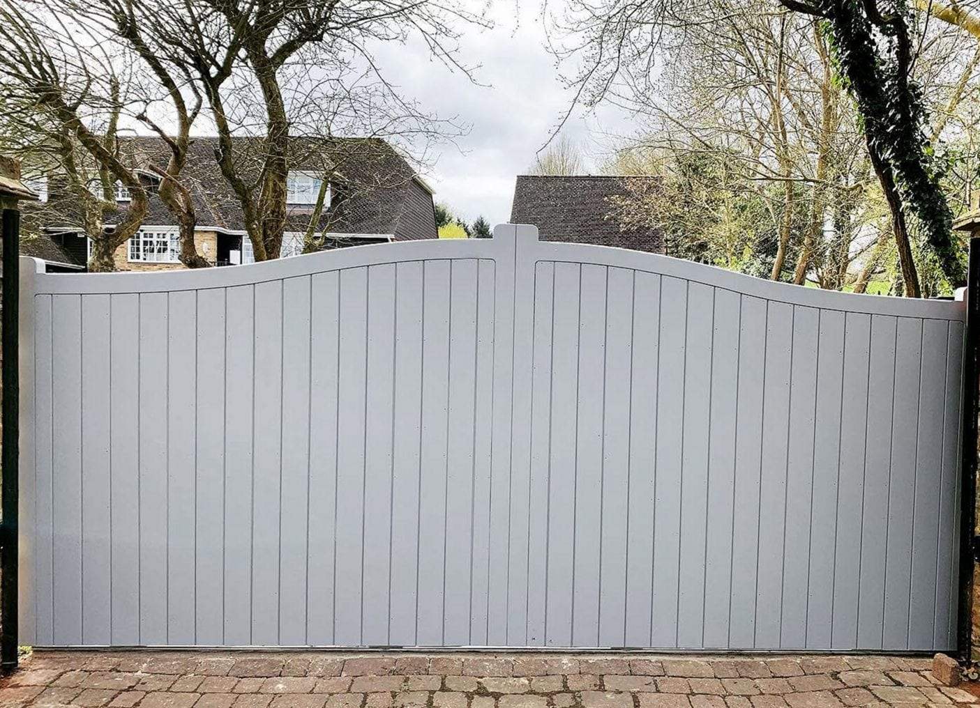 Light gray painted timber gate with vertical boards and a swept, bell-curve top across a block-paved driveway, with bare trees and houses behind