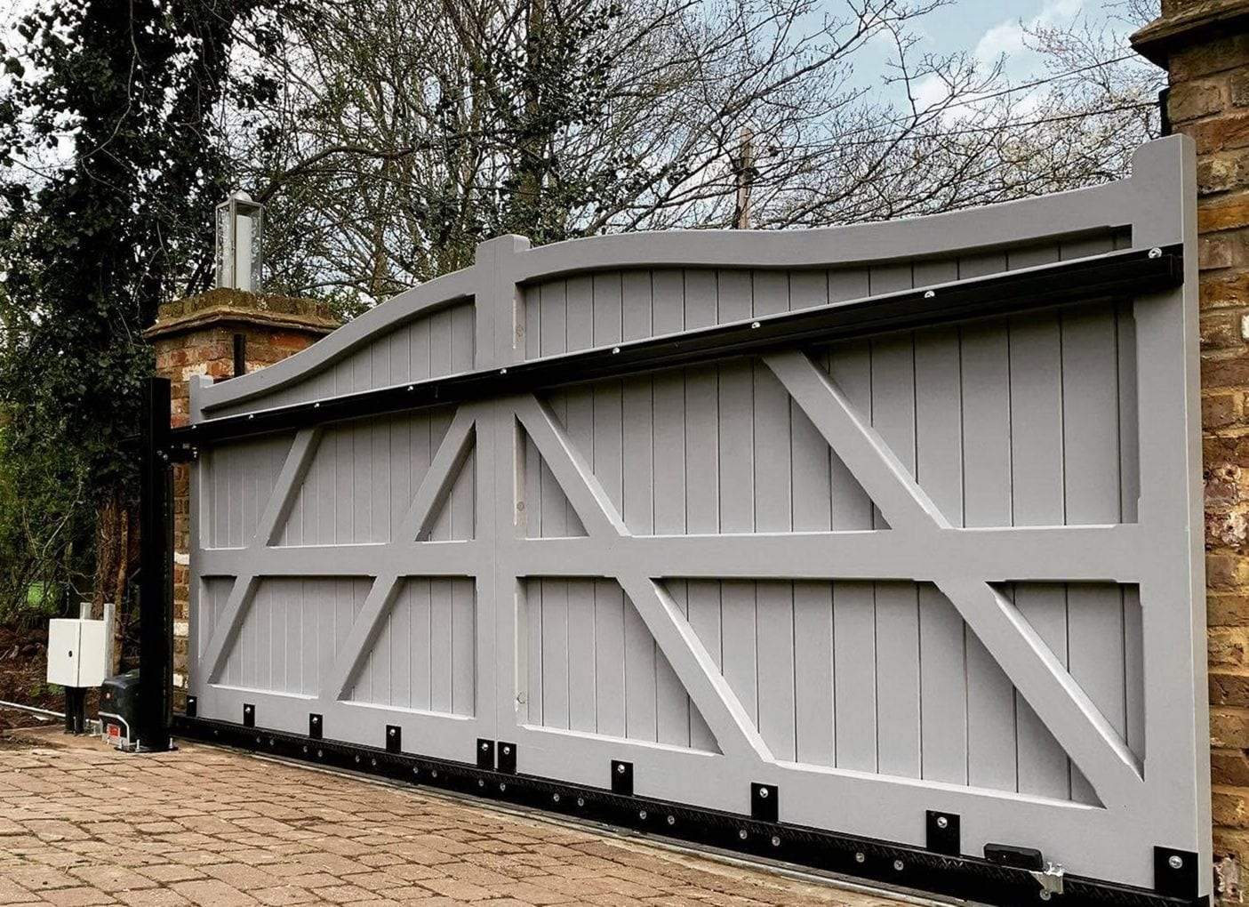 Back side of a gray painted timber slide gate showing its Z-shaped bracing, a toothed gear rack bolted along the bottom rail, the gate operator and a gray control box at the left end beside a brick pier