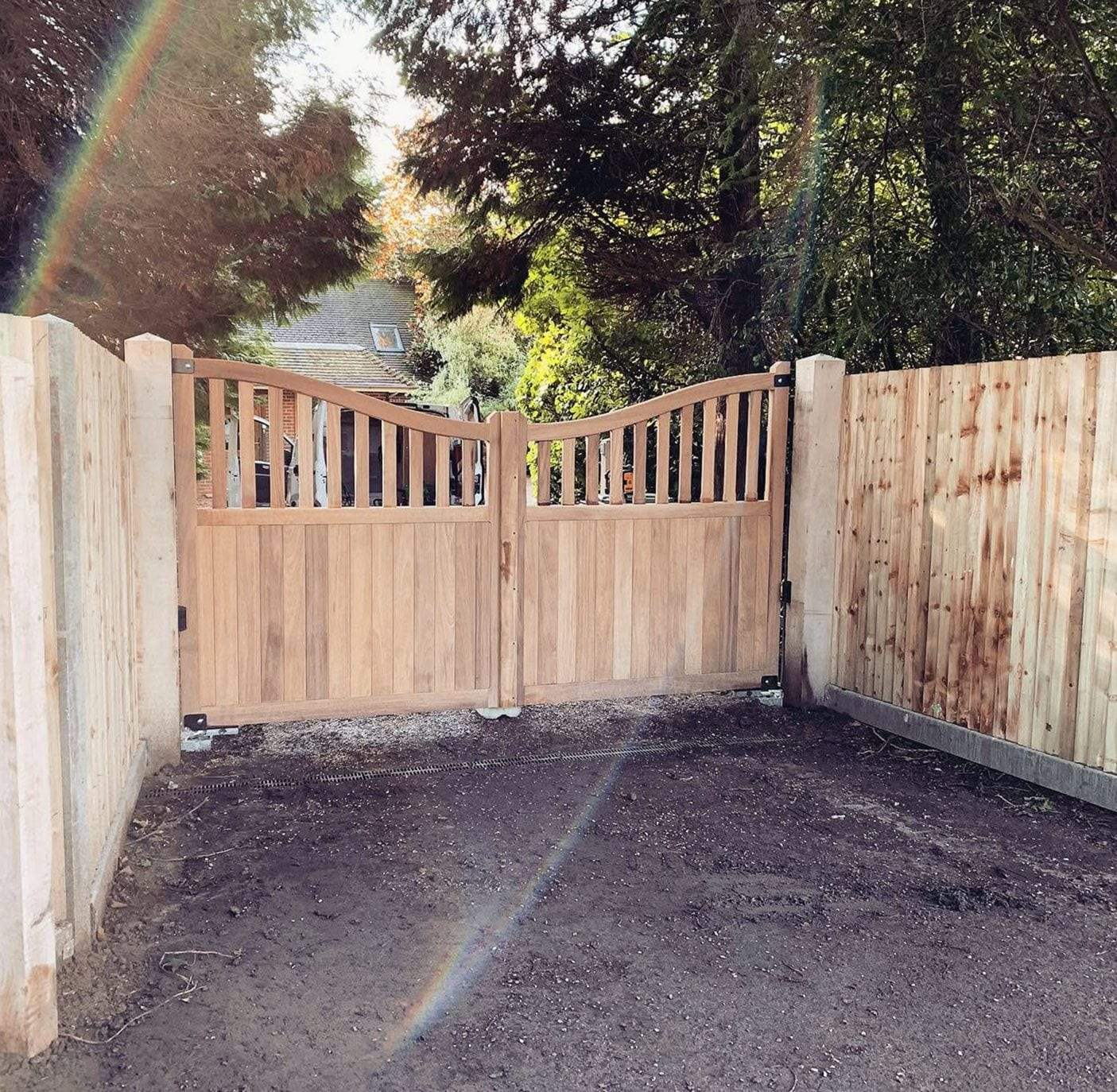 Newly installed pair of natural hardwood swing gates with an open slatted top in a gentle curve, between new square posts and new board fencing, across a bare soil driveway with a drainage channel